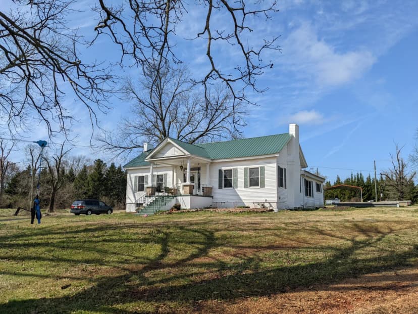 The 1832 farmhouse at 5400 Ridgewood Ave, Anniston, Alabama — white clapboard with green metal roof, set in an open field in winter, February 2022