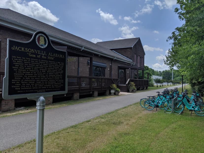 The renovated Jacksonville train station on the Chief Ladiga Trail with a historical marker for Jacksonville, Alabama and teal bike share bikes out front, 2019