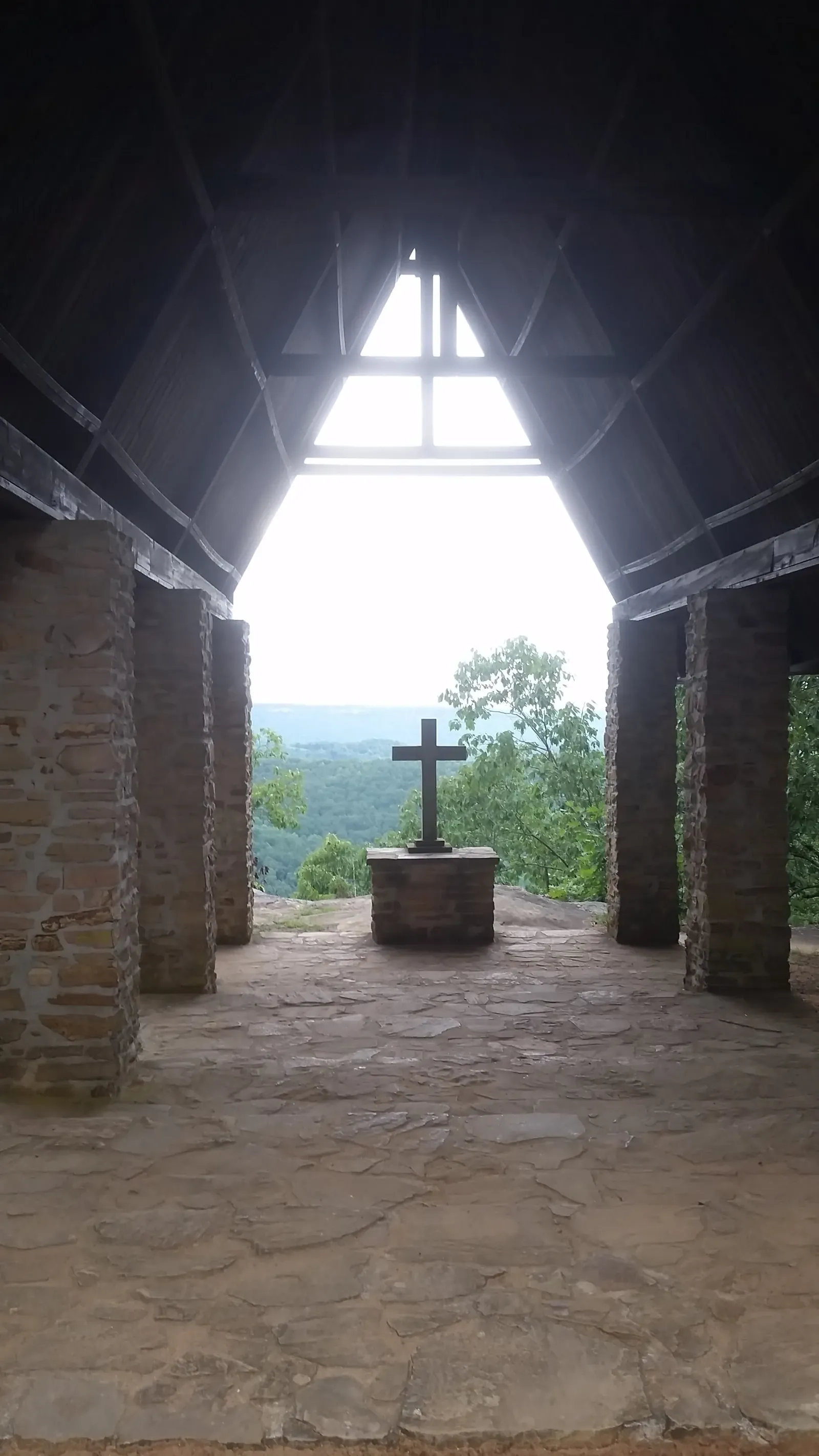 Interior of the open-air Chapel on the Mountain at Camp Sumatanga, a stone cross on a pedestal framed by the open triangular gable, the St. Clair County valley falling away beyond it