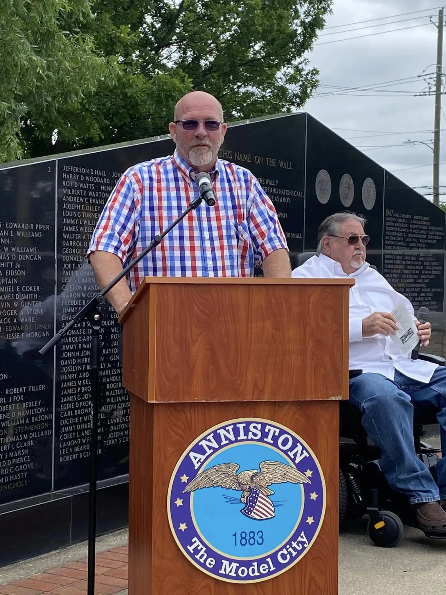 Jay Jenkins speaking at the Anniston Veterans Memorial podium, names on the memorial wall behind him, May 2023