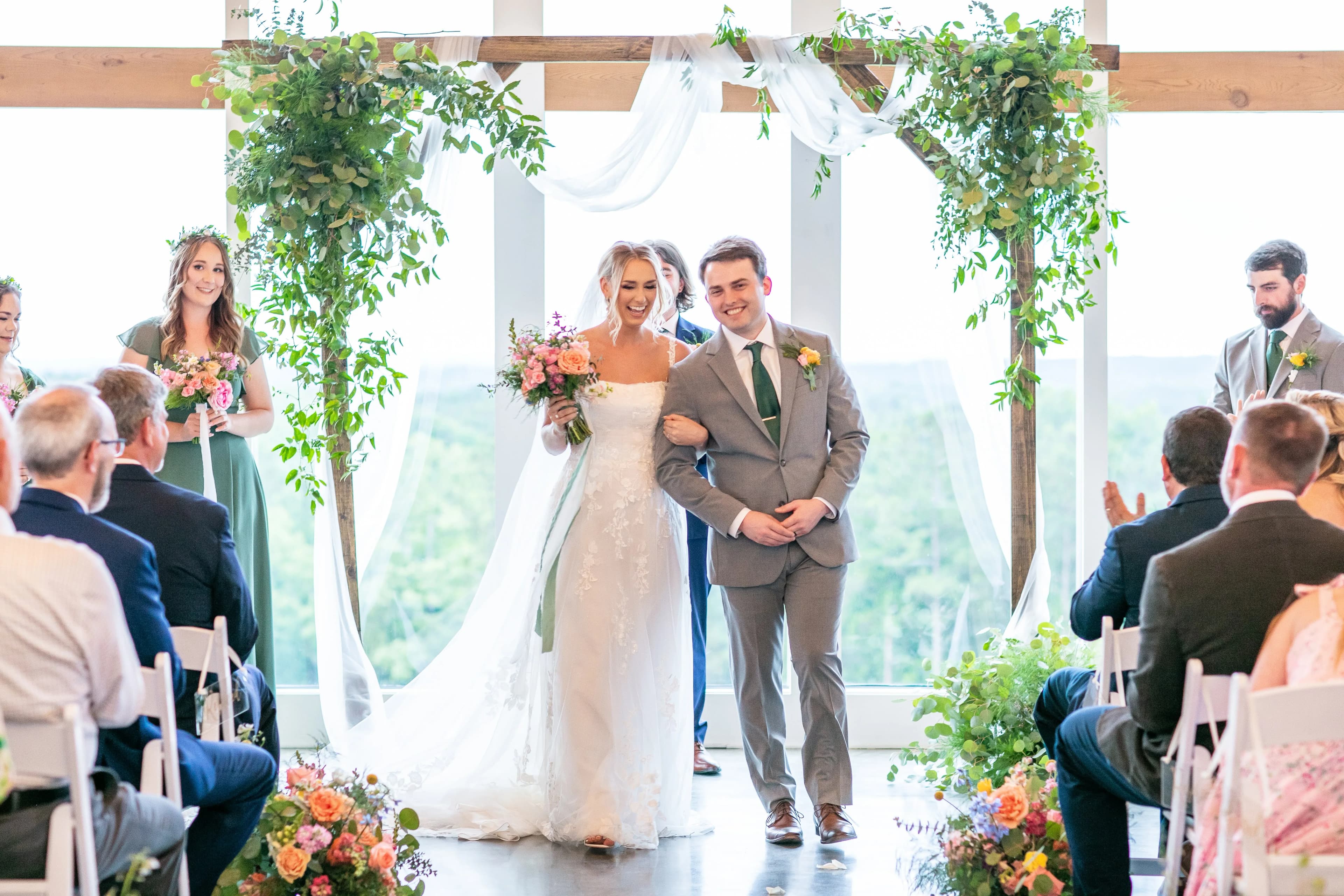 Sonny Jenkins and Sam walking back down the aisle at the wedding ceremony, a wooden arch with greenery behind them, panoramic windows opening to green hills