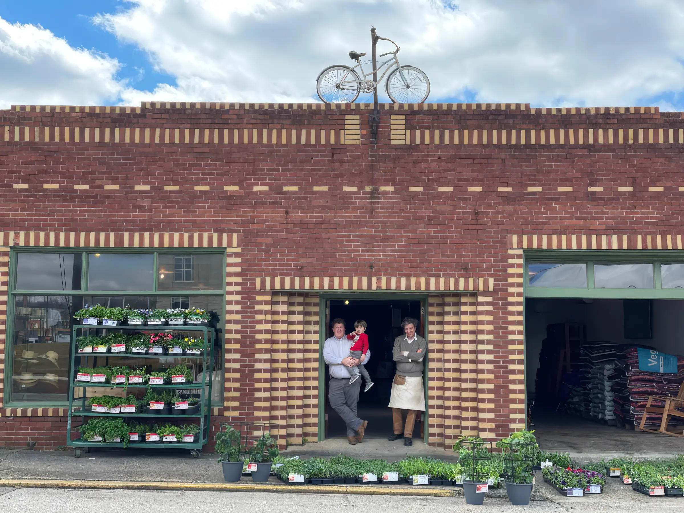 Lewis Downing and his father RD standing in the doorway of Downing and Sons on Gurnee Avenue in Anniston, with spring plants displayed on the sidewalk and a bicycle mounted above the entrance