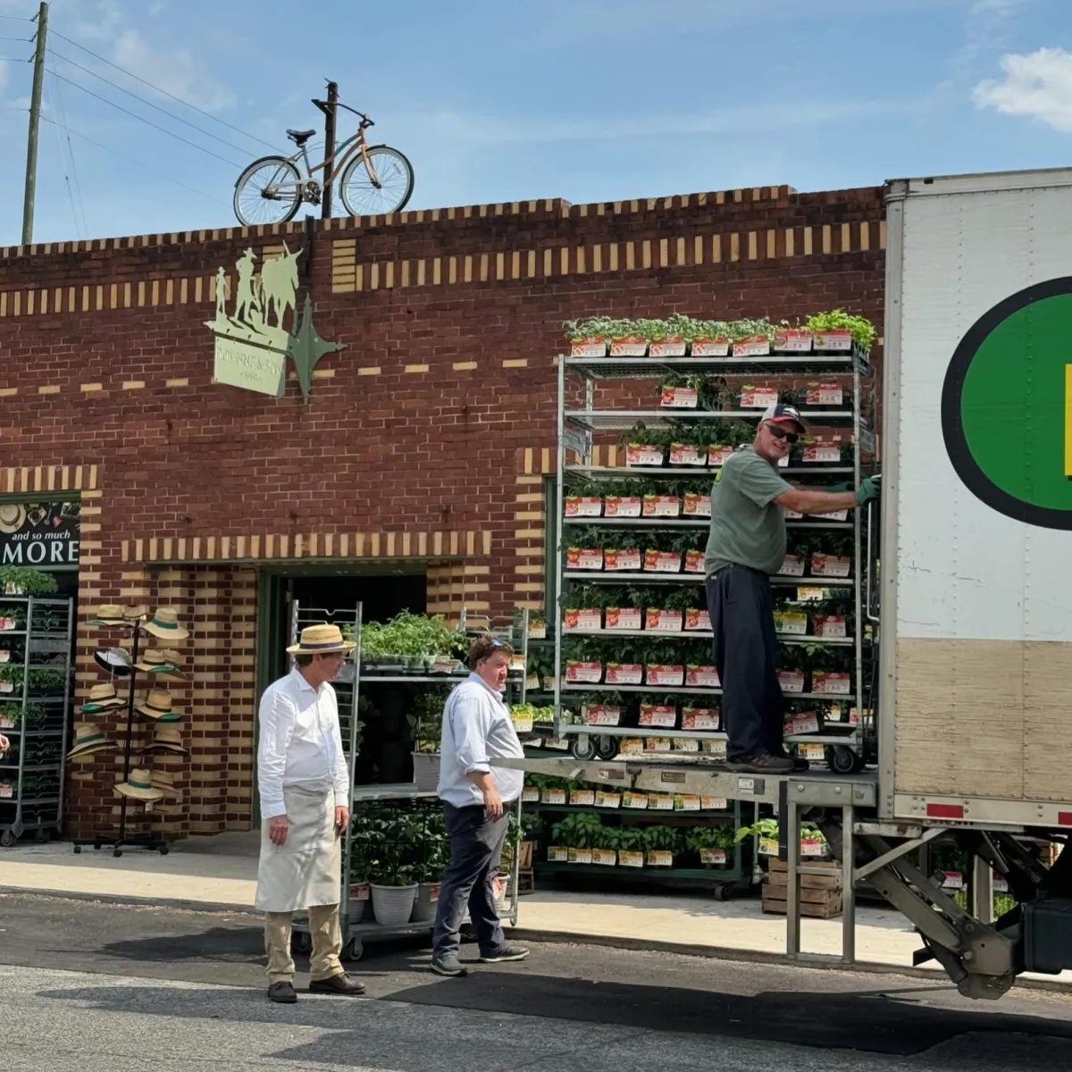 RD Downing in a straw hat and apron standing outside with Lewis as workers unload a rack of potted plants from a delivery truck in front of Downing and Sons on Gurnee Avenue