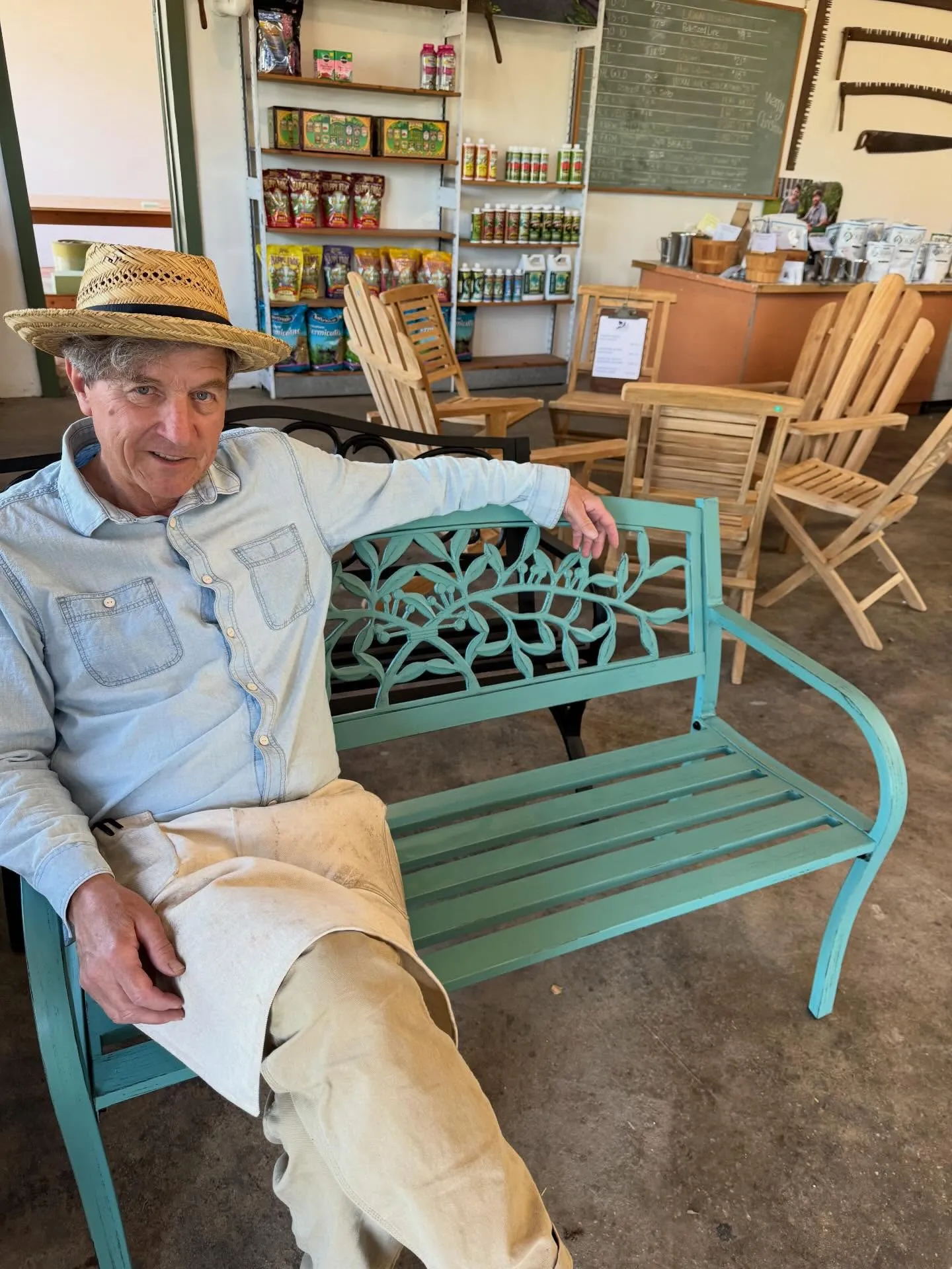RD Downing in a straw hat sitting on a teal garden bench inside Downing and Sons, surrounded by store inventory