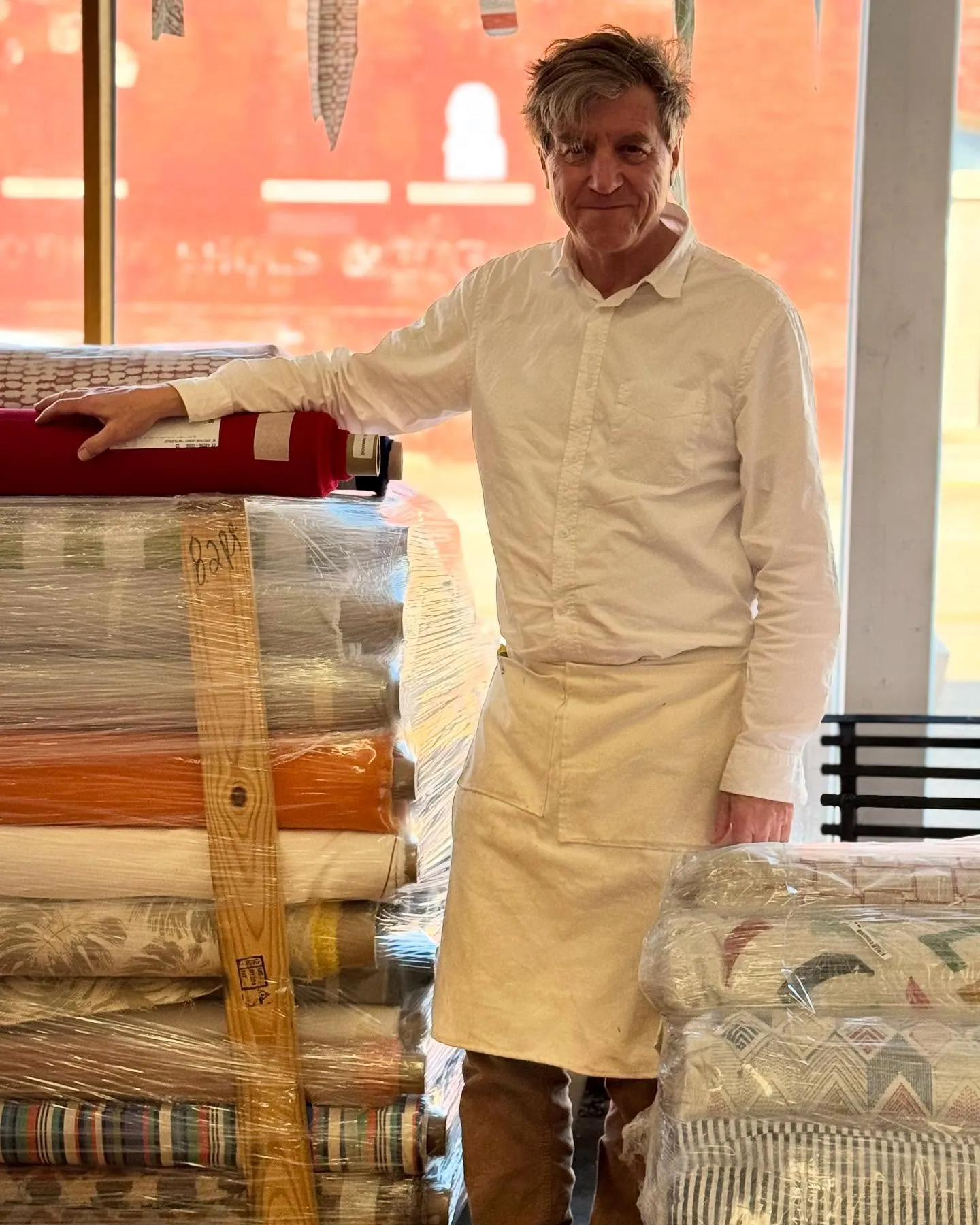 RD Downing in his work apron standing among stacked bolts of upholstery fabric at Downing and Sons