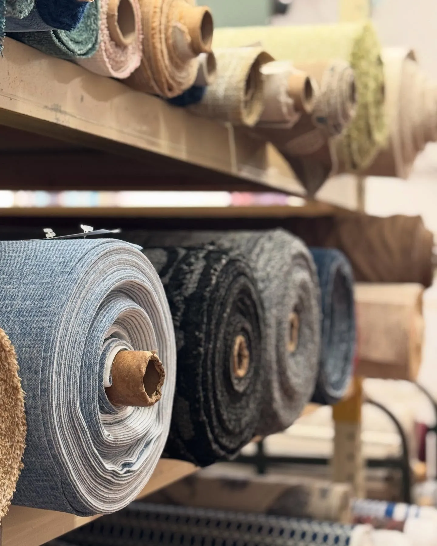 Rolls of upholstery fabric stacked on shelving at Downing and Sons, including blues, grays, tans, and textured wovens